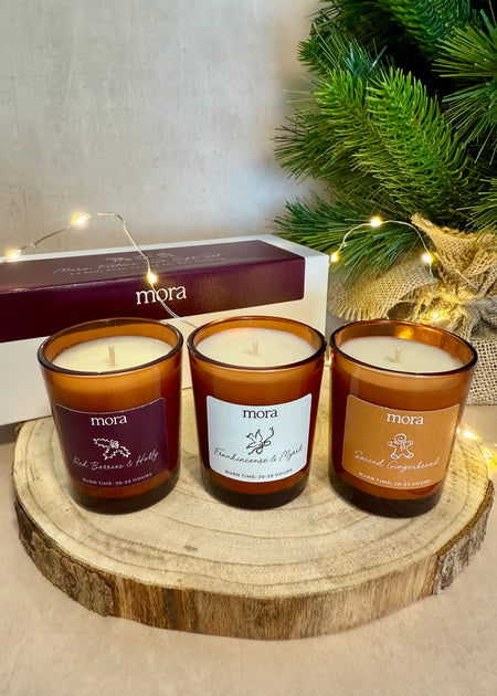 Votive gift set. Three 'mora' candles on a wooden stand with a Christmas tree in the background. Amber jars. 