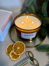 Lit candle with 'mora' label on a surface with dried oranges and greenery