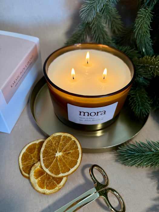 Lit candle with 'mora' label on a surface with dried oranges and greenery