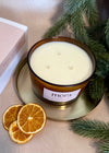Three-wick candle labeled 'mora' on a gold tray with dried oranges and greenery. Christmas collection