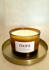 Amber glass jar candle with 'mora' label on a gold tray against a beige background. three cotton wicks. soy wax. 