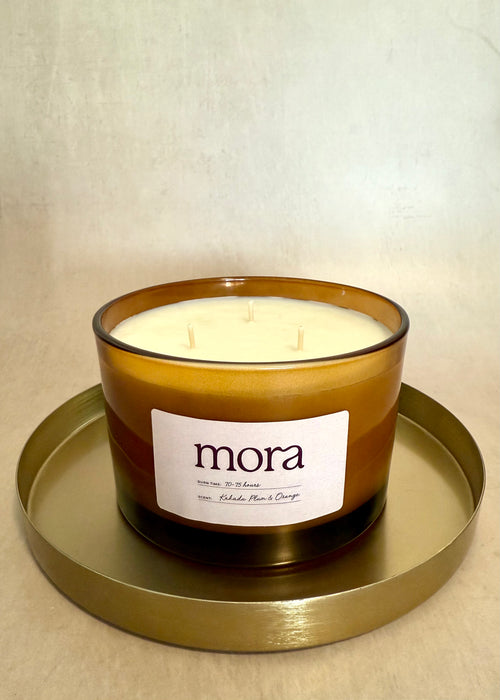 Amber glass jar candle with 'mora' label on a gold tray against a beige background. three cotton wicks. soy wax. 