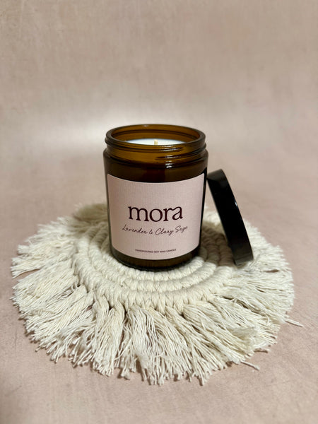 Candle in a brown jar labeled 'mora' on a beige surface with a textured mat.