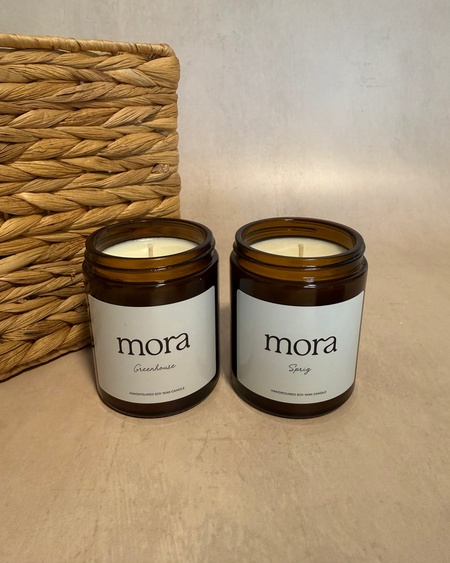 Two 'mora' candles on a beige surface with a woven basket in the background.