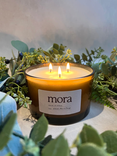 A lit 3-wick candle with a mora label in an amber glass jar, surrounded by green foliage. christmas scene