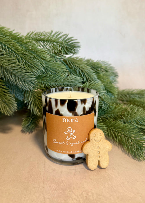 Candle with a gingerbread man and 'mora' label next to greenery on a beige surface christmas foliage