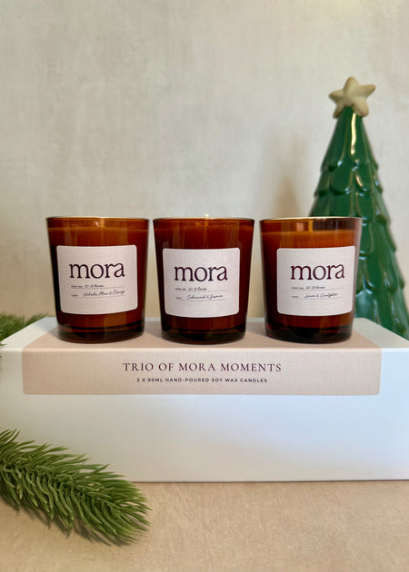 Three 'mora' candles on a white surface with a decorative tree in the background.