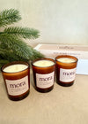 Three 'mora' candles on a beige surface with a green branch and product packaging in the background.